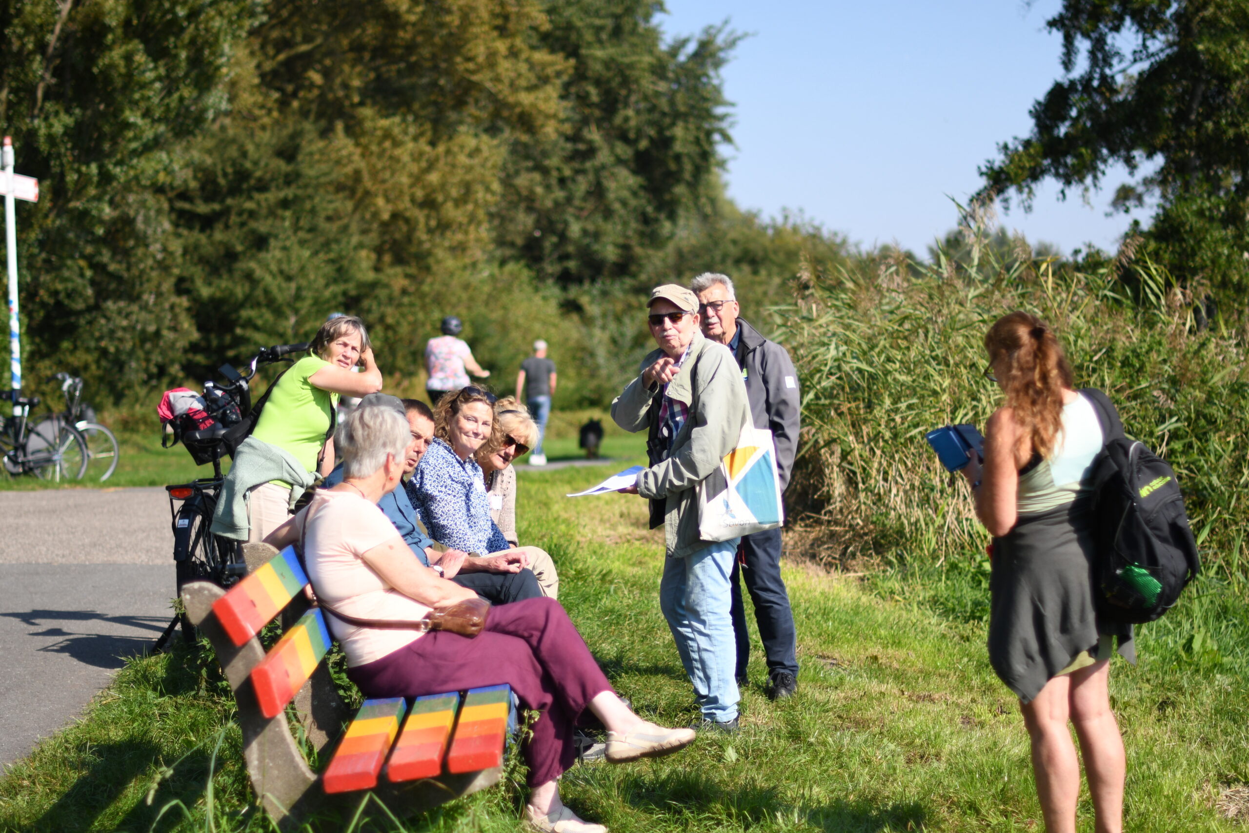 Groep mensen rusten op een kleurrijke bank langs een pad in een groen landschap.