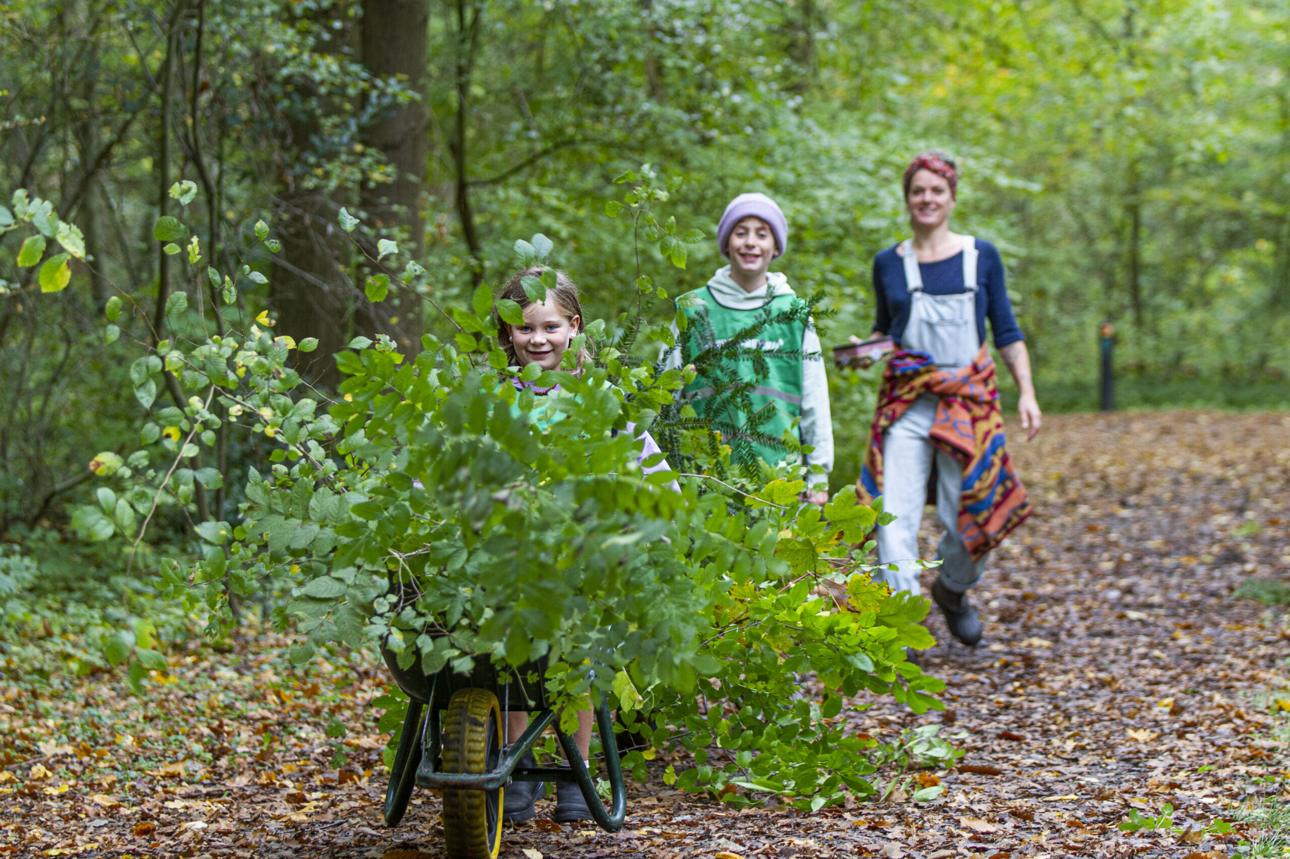 Kinderen duwen een kruiwagen vol bladeren in een bos, gevolgd door een volwassen vrouw.