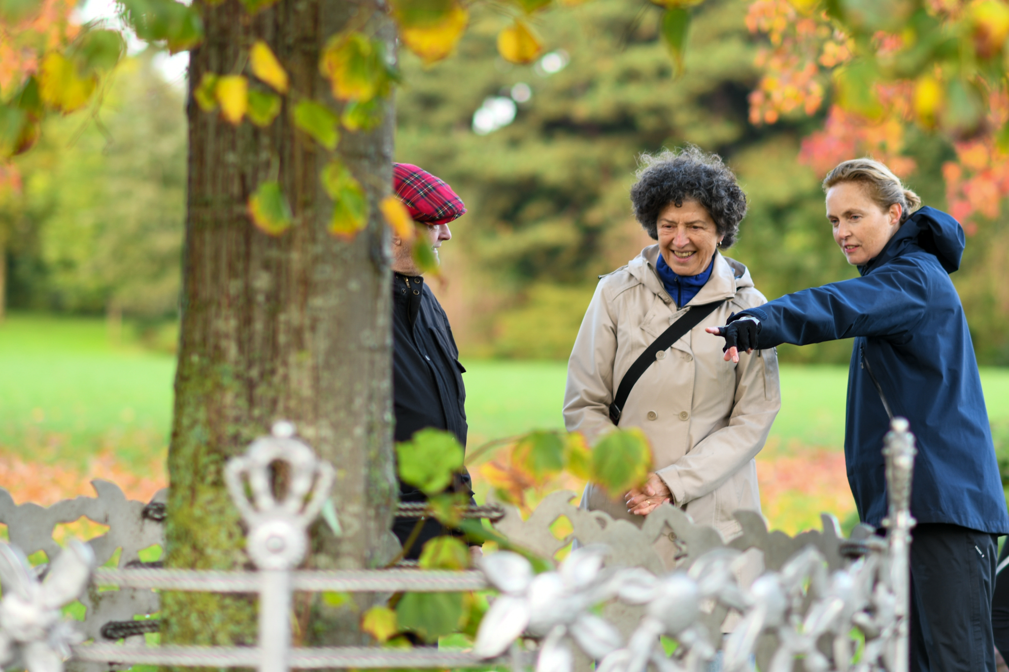 Drie mensen in park, vrouw in blauwe jas wijst iets aan. Herfstbladeren op de achtergrond.