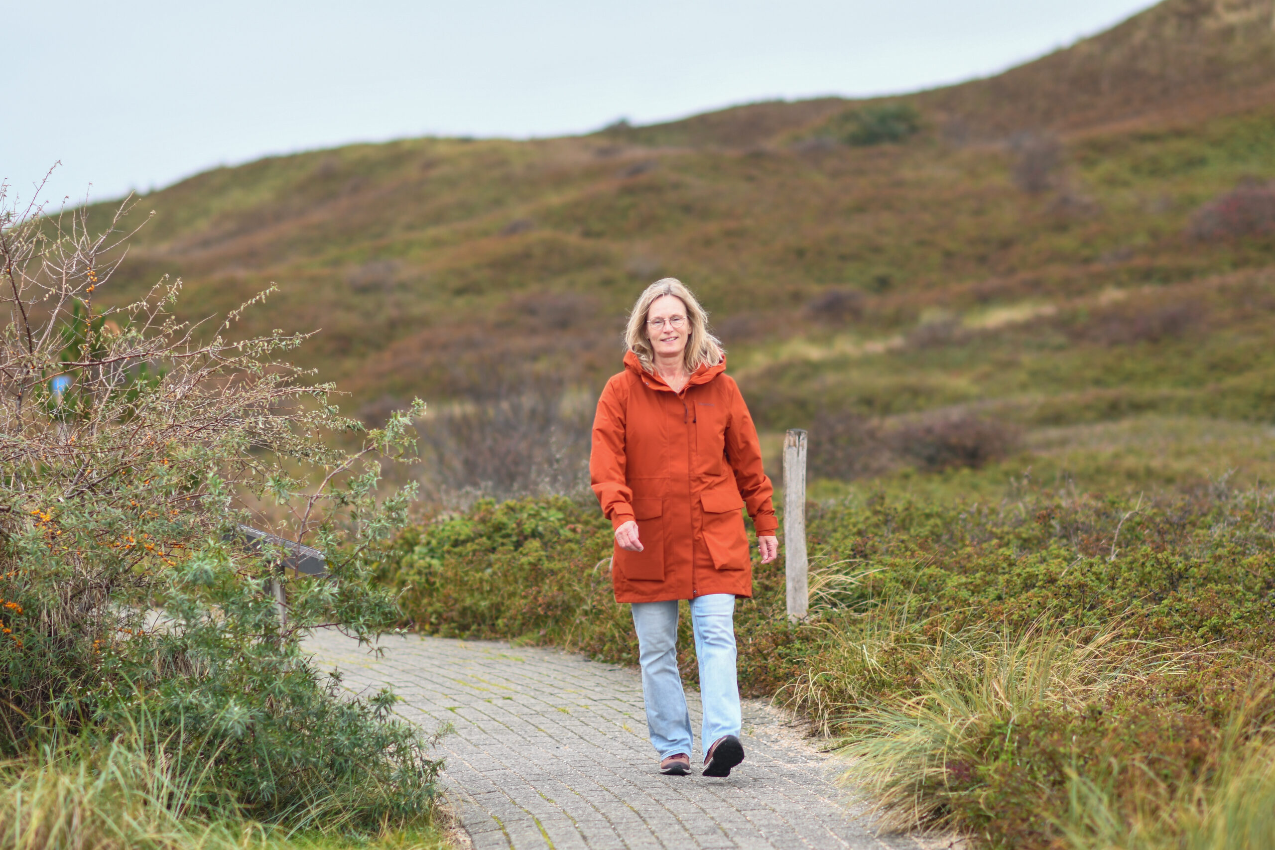 Vrouw in oranje jas wandelt op een pad door groene duinen.