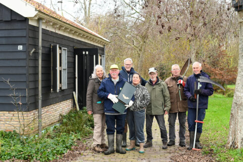 Groep mensen poseert voor donker gebouw met gereedschap in een tuinomgeving.
