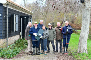 Groep mensen voor een houten huisje met tuingereedschap en een vleermuizenhuis, omgeven door bomen.