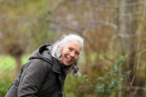 Oudere vrouw met grijs haar in jas glimlacht in een bosrijke omgeving.