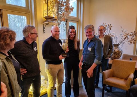 Een groep mensen viert een prijsuitreiking binnenshuis, met een trofee en glimlachende gezichten.