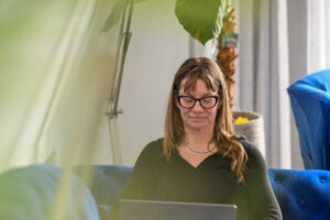 Vrouw met bril zit op blauwe bank en werkt geconcentreerd op laptop, omringd door planten.