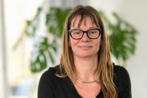 Vrouw met bril en bruine haren tegen een groene, wazige achtergrond binnen.