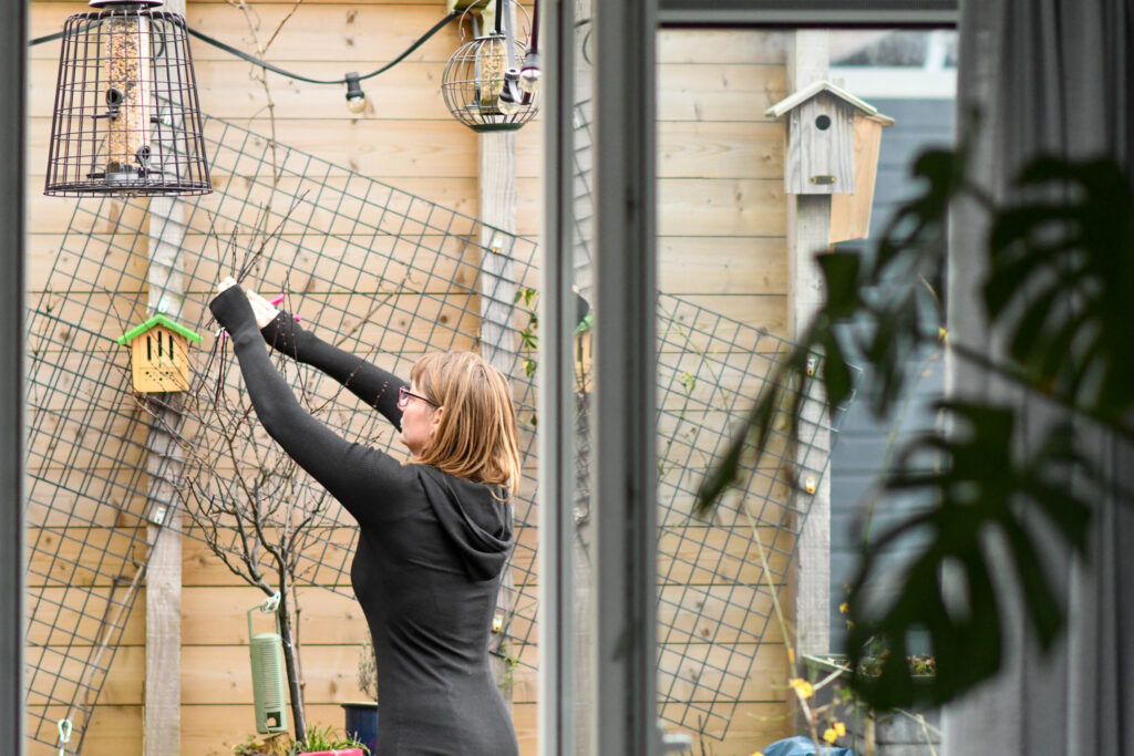 Vrouw in tuin hangt vogelvoeder, houten wand met nestkastjes en gaas zichtbaar.