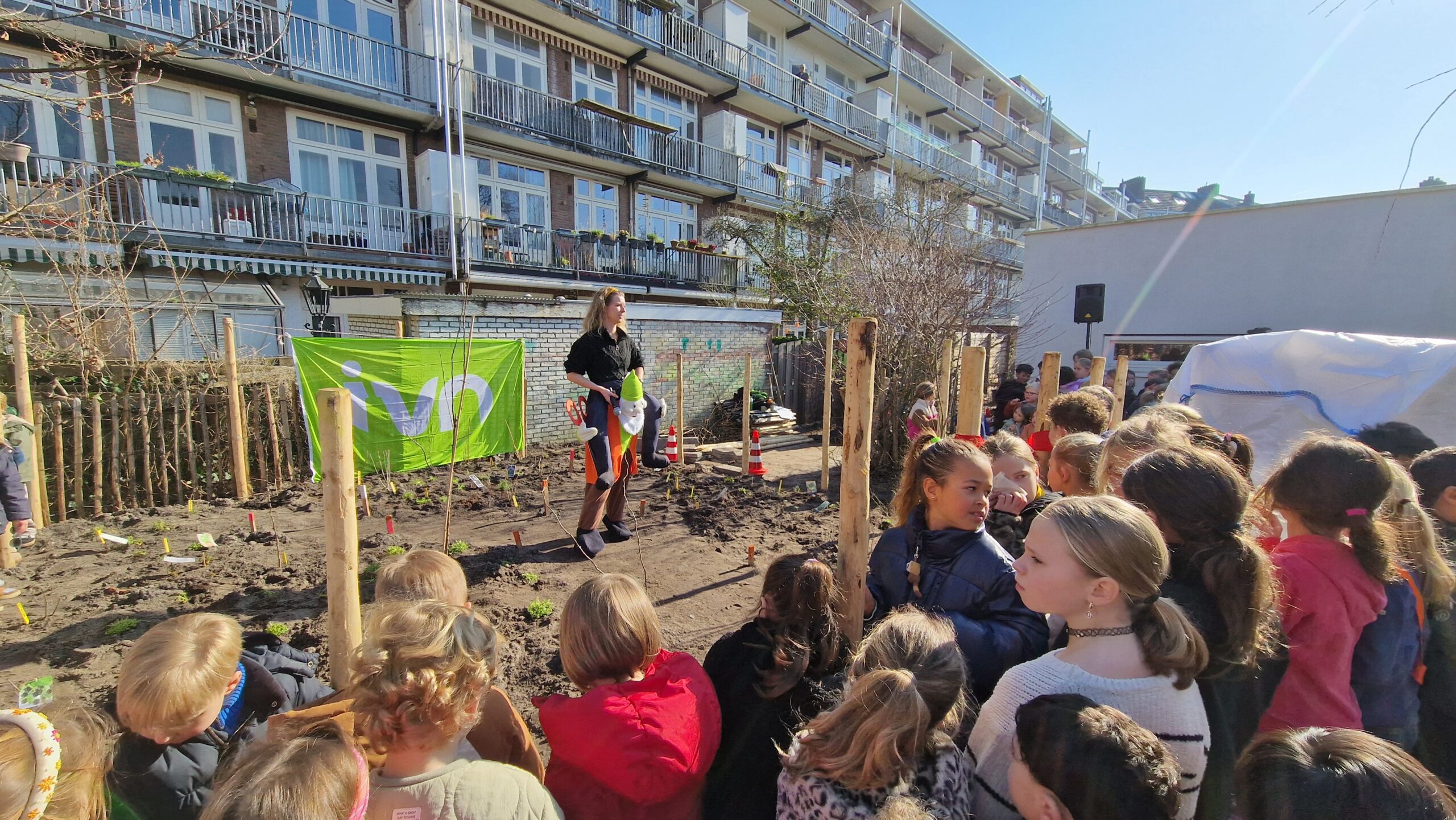Vrouw spreekt voor groep kinderen bij gemeenschappelijke tuin met groene banner en flats op achtergrond.