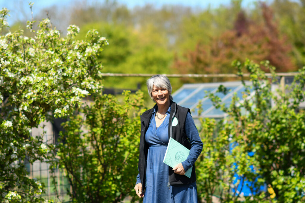 Vrouw in blauwe jurk en zwarte vest staat glimlachend in een tuin met groene planten en bloesembomen.