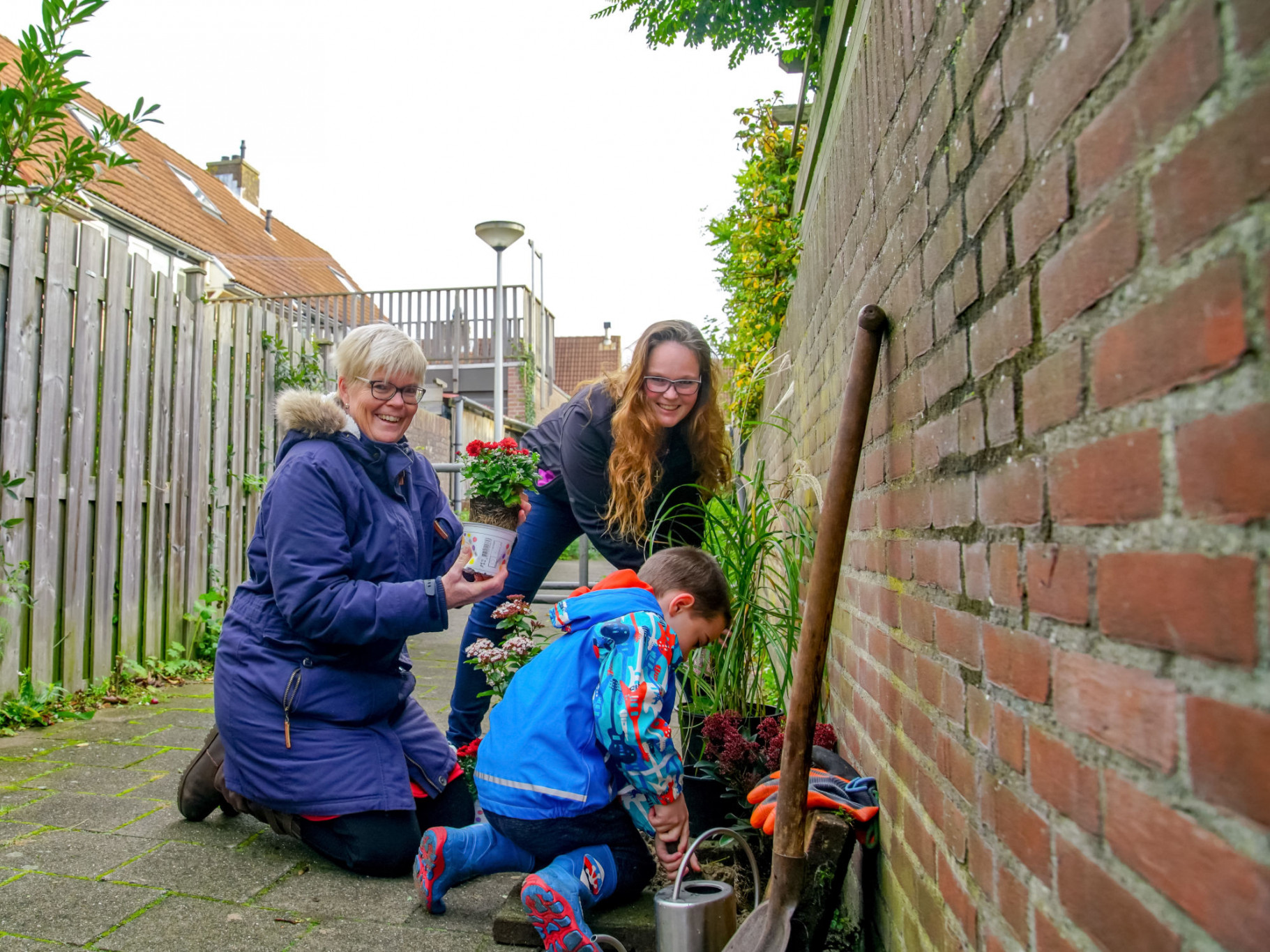 Mensen planten bloemen in een smalle tuin, naast een bakstenen muur en houten schutting.