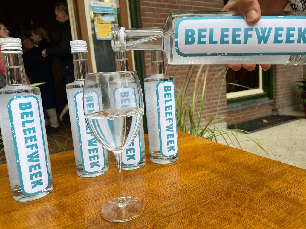 Fles met label "Beleefweek" schenkt drank in een glas; meer flessen op houten tafel.