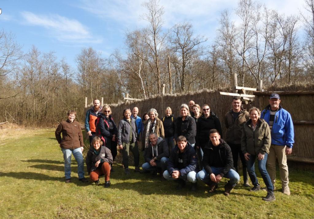 Groep mensen poseert buiten voor een natuurlijke afscheiding, omgeven door bomen en gras.