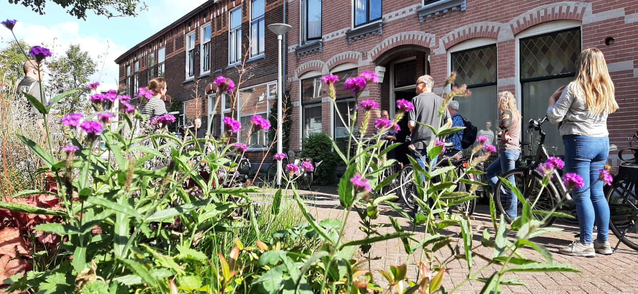 Groep mensen voor een rij bakstenen huizen, bloemen op de voorgrond.