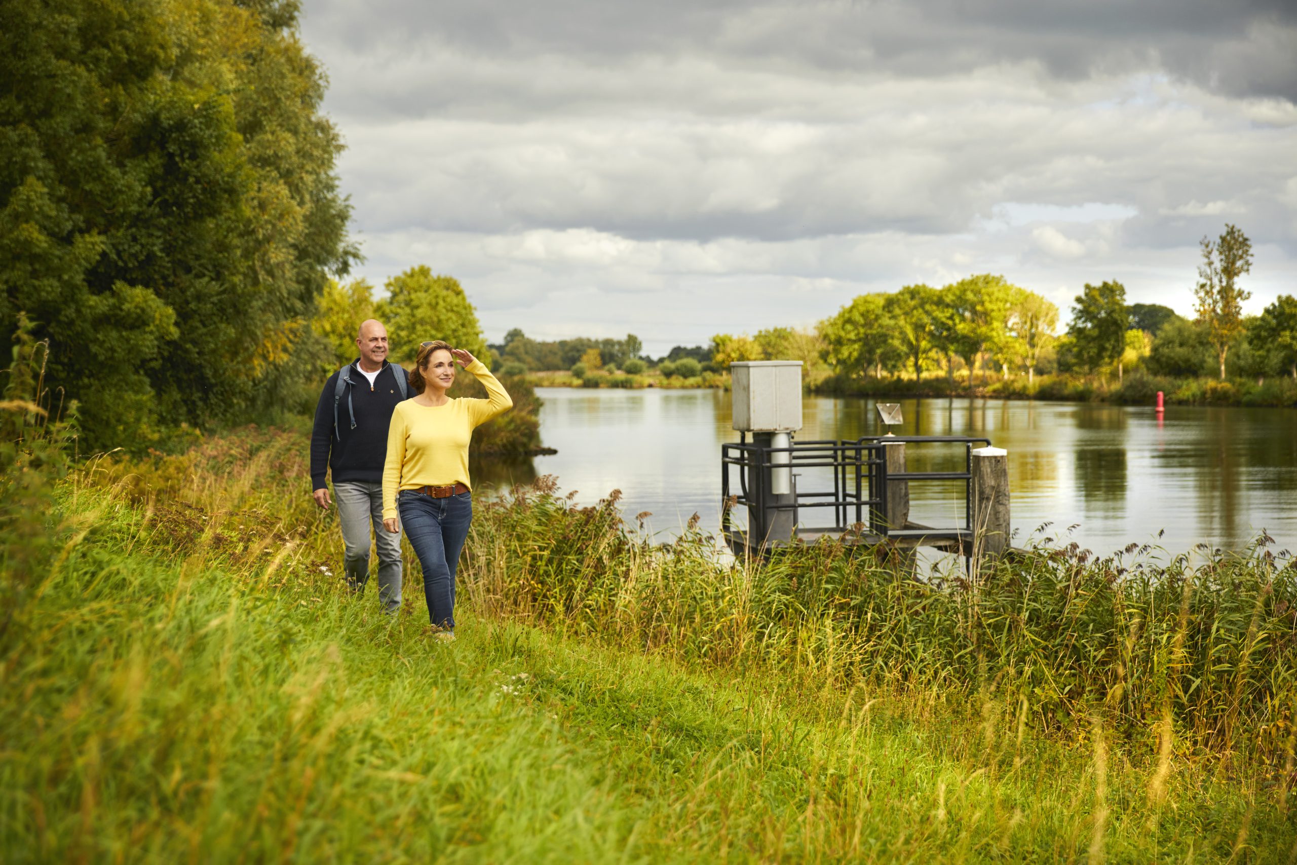 Een man en vrouw wandelen langs een rivier omgeven door groene natuur onder een bewolkte hemel.