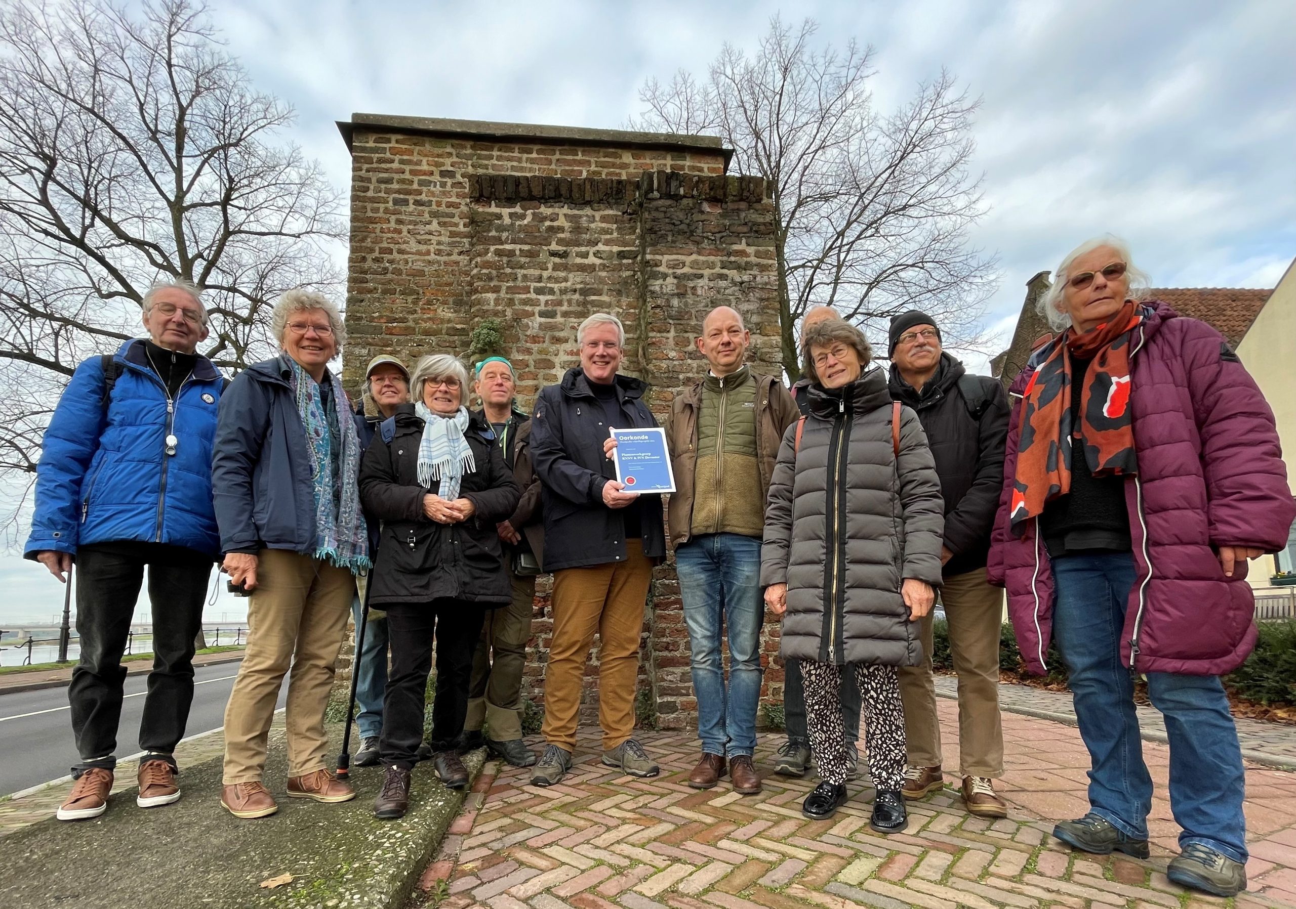 Groep mensen poseert bij een oude bakstenen muur, met een certificaat in het midden.
