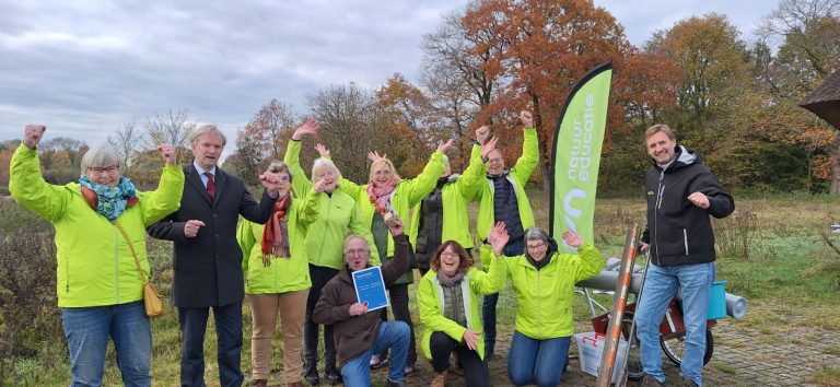 Groep mensen in groene jassen viert buiten met certificaat, naast een 
