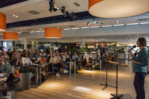 Een vrouw spreekt met een microfoon voor een zittend publiek in een verlichte, moderne zaal.