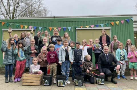 Groep kinderen en een volwassene poseren buiten voor een groene muur met kleurrijke vlaggetjes.