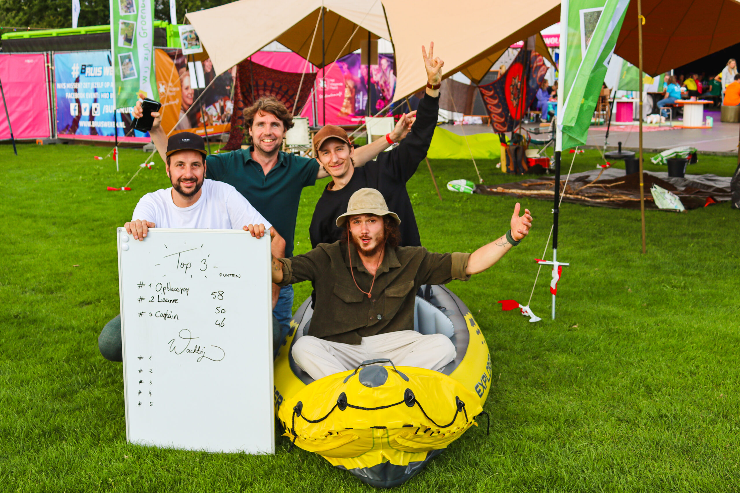 Vier mannen poseren met een bord dat een top 3 ranglijst toont op een grasveld bij een kleurrijke tent.