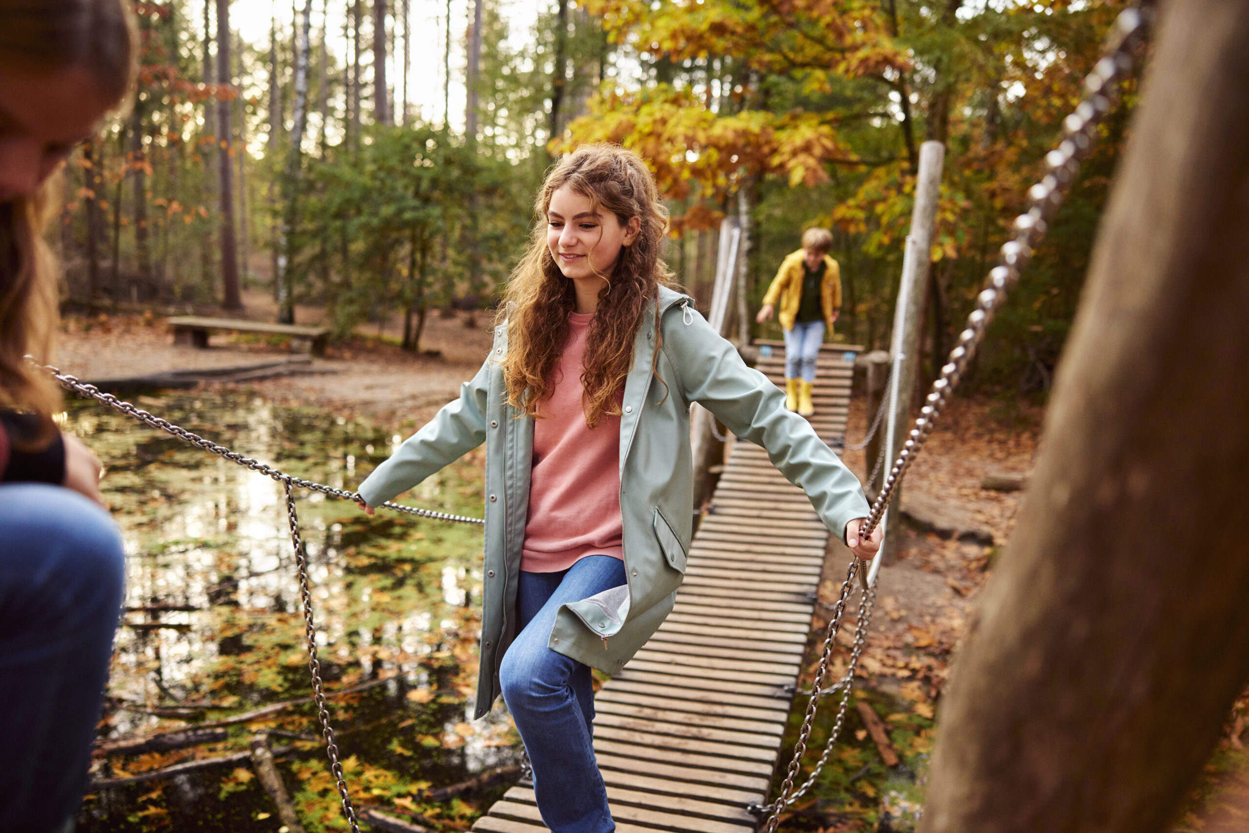 Meisje loopt over een touwbrug in een herfstbos met een vijver eronder.