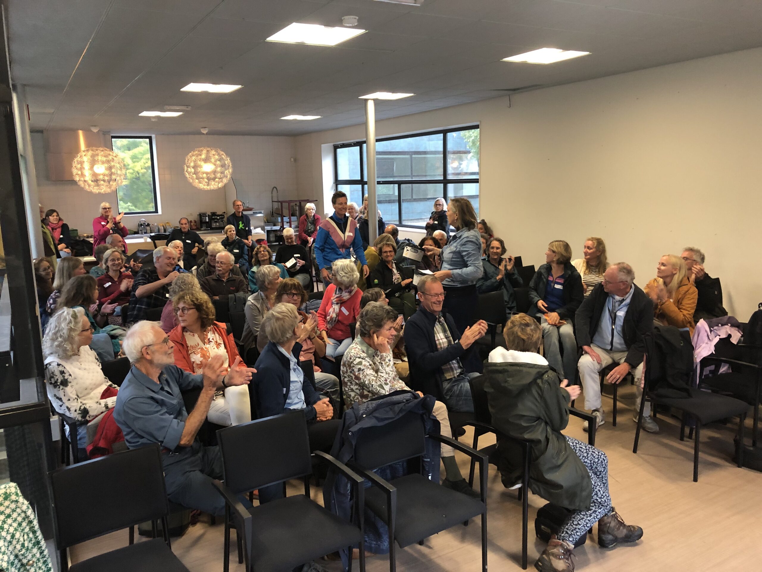 Een groep mensen applaudisseert en luistert tijdens een bijeenkomst in een zaal.