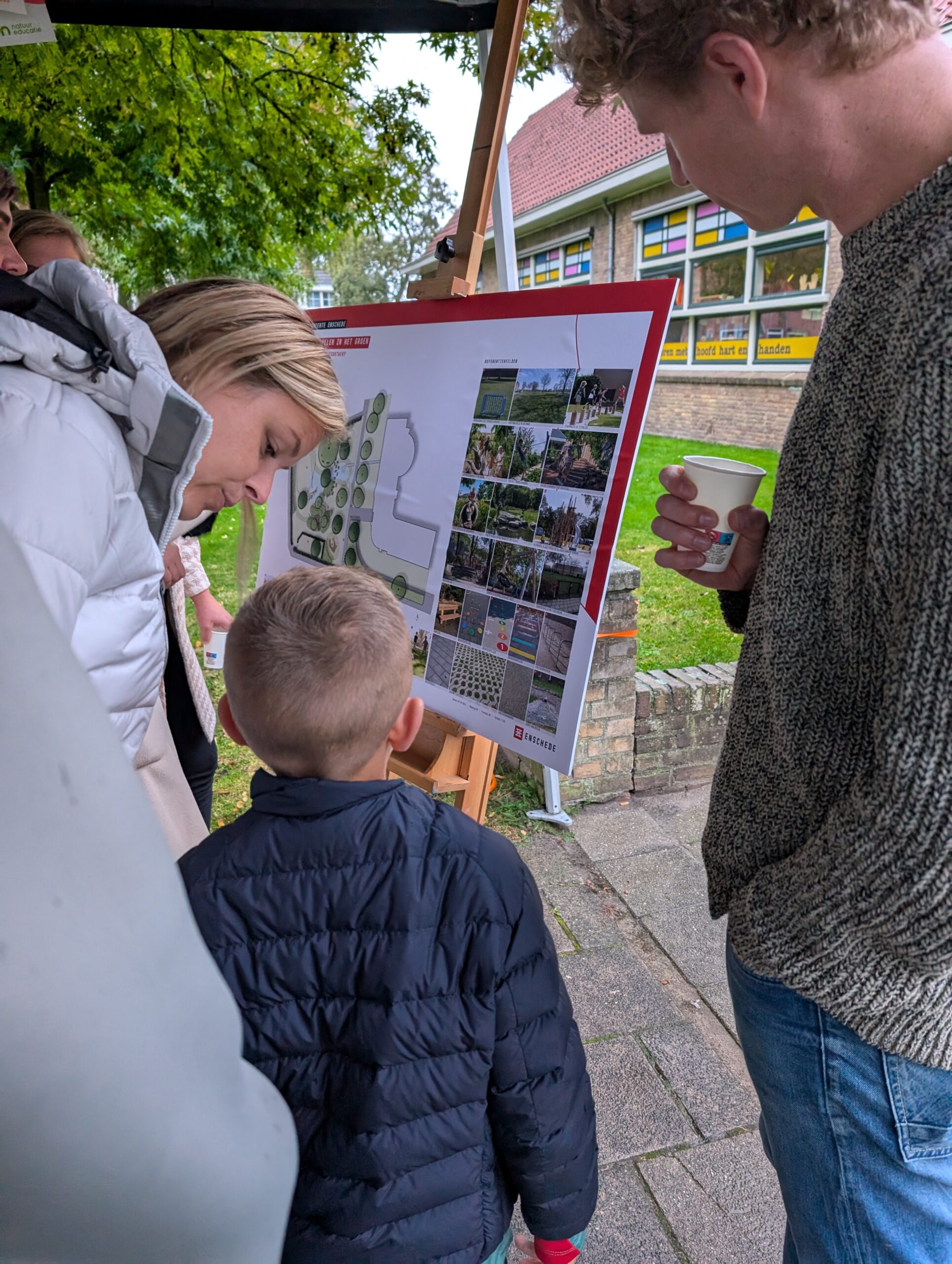 Mensen bekijken een bord met ontwerpen en foto's bij een scholengebouw in een groene omgeving.