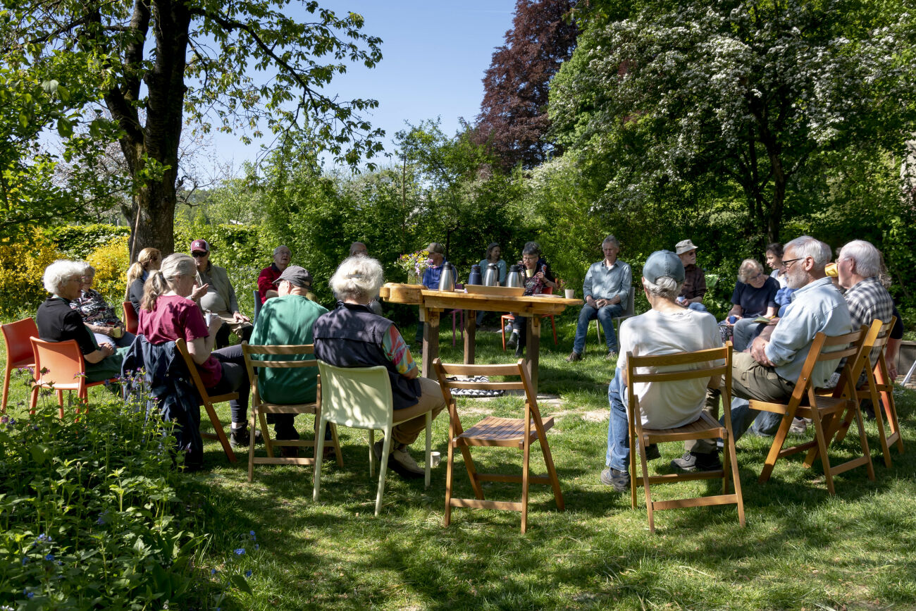 Een groep mensen zit in een kring op stoelen in een tuin, omringd door bomen en struiken.