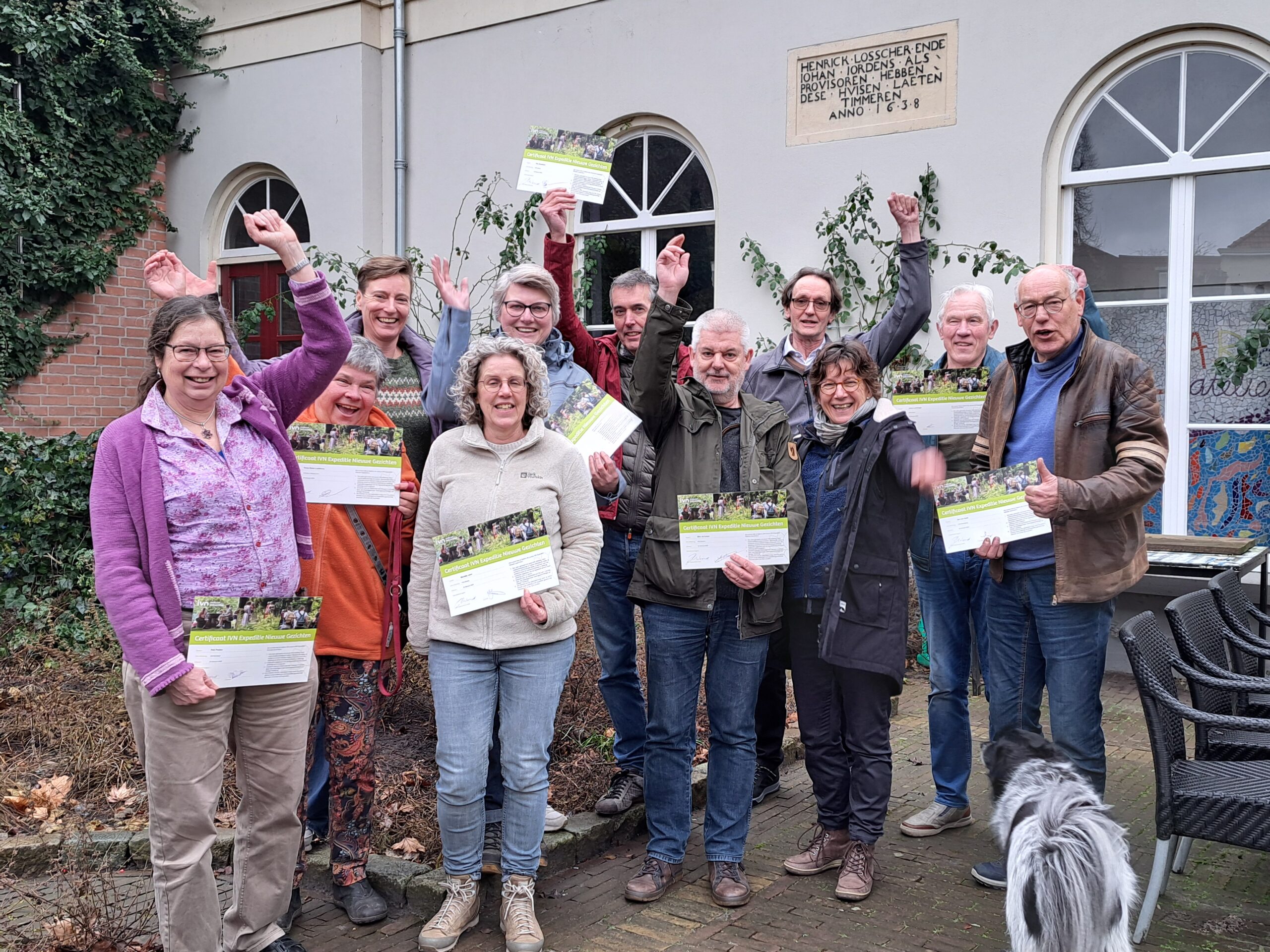 Groep mensen juicht en toont certificaten voor gebouw, naast een zwart-witte hond.