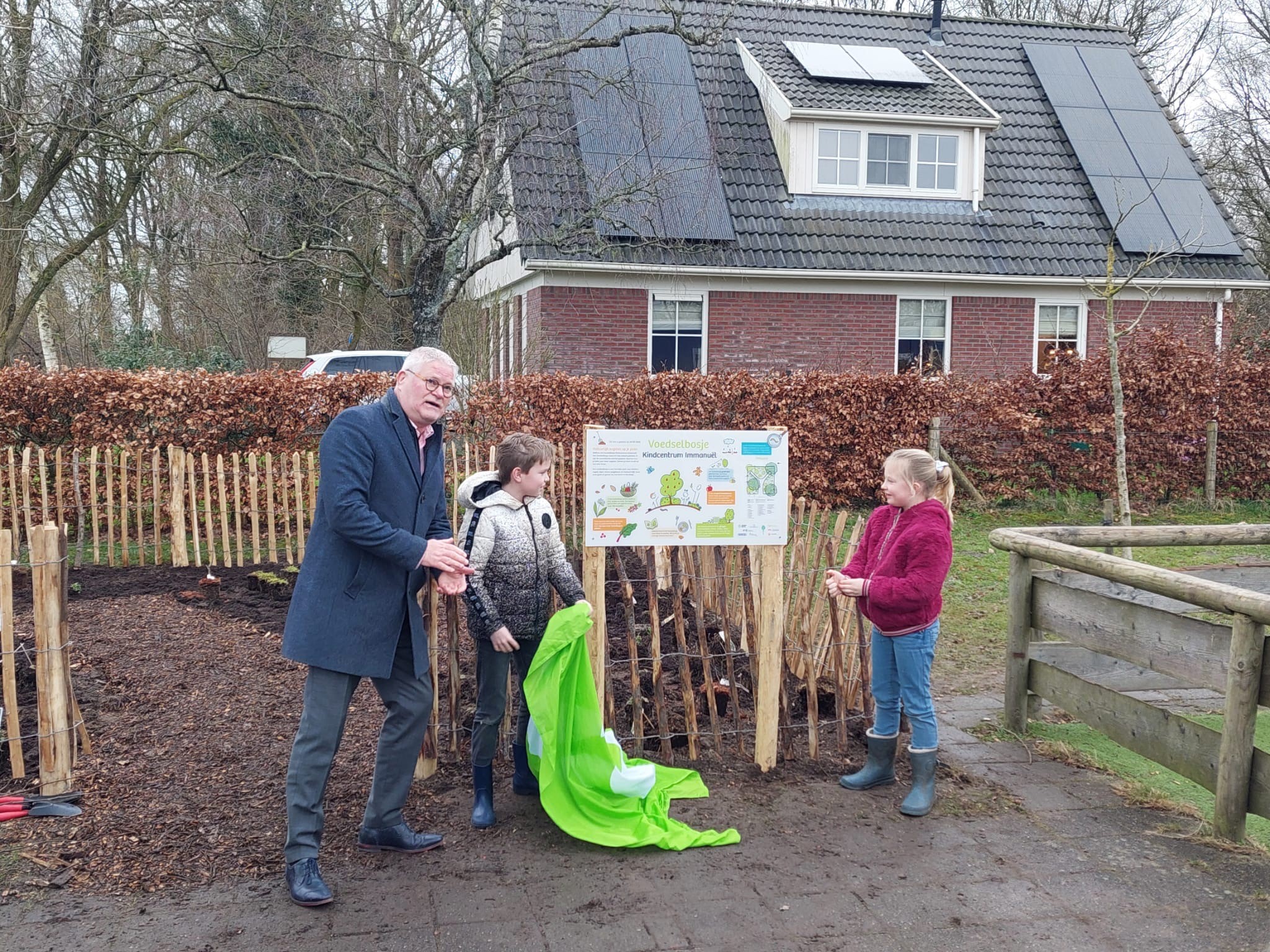 Man en twee kinderen onthullen informatiebord bij een tuin. Houten hek en huis op achtergrond.