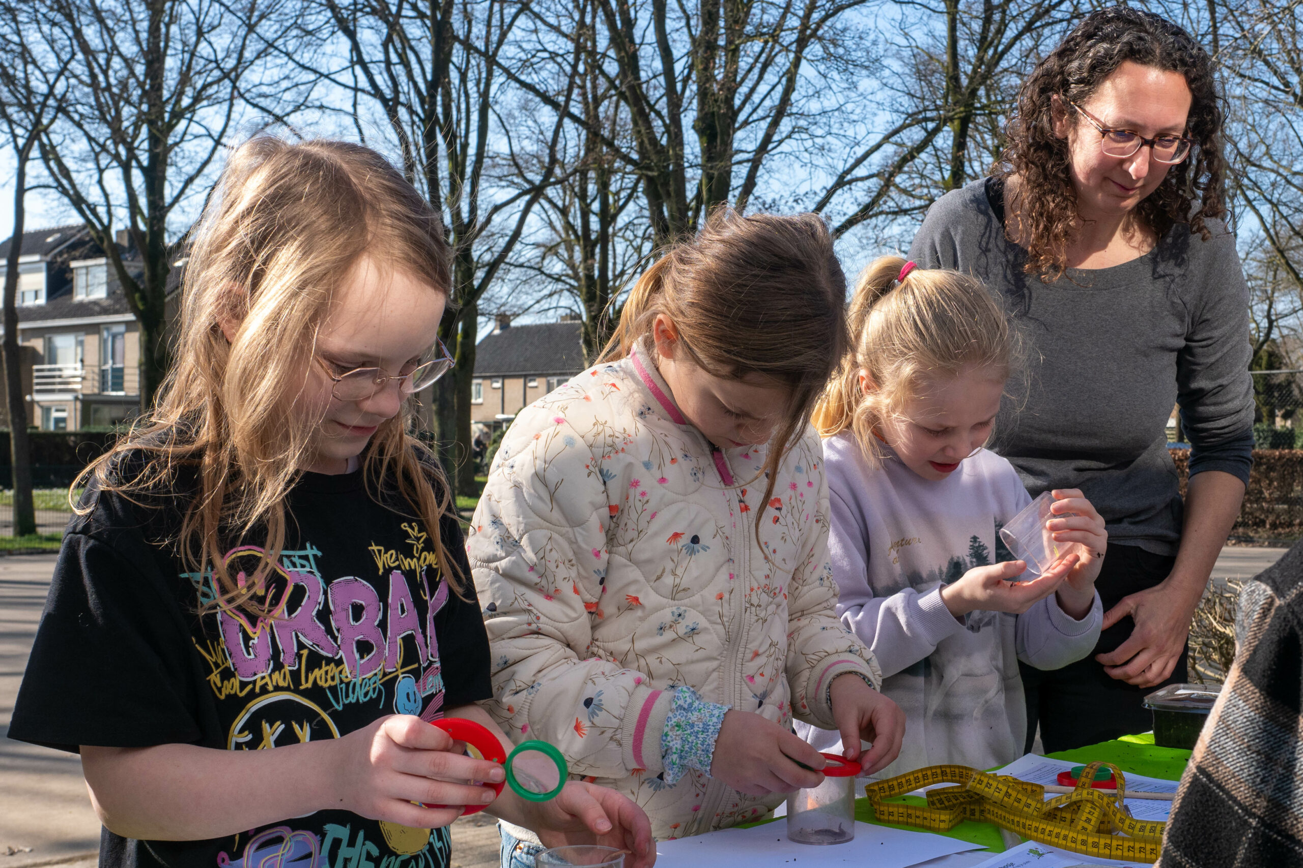 Drie kinderen en een volwassene onderzoeken potjes buiten op een zonnige dag.