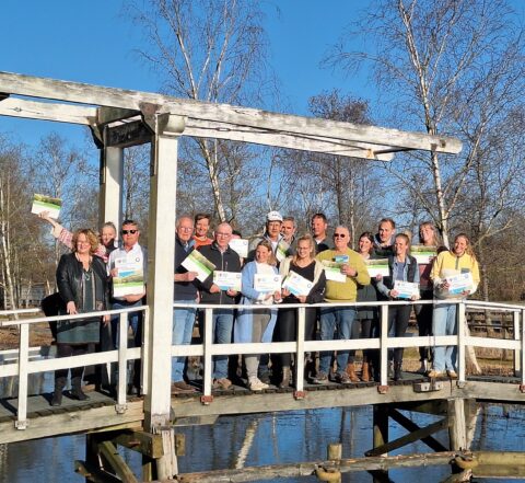Groep mensen op een houten brug, poseren met certificaten onder een heldere, blauwe lucht.