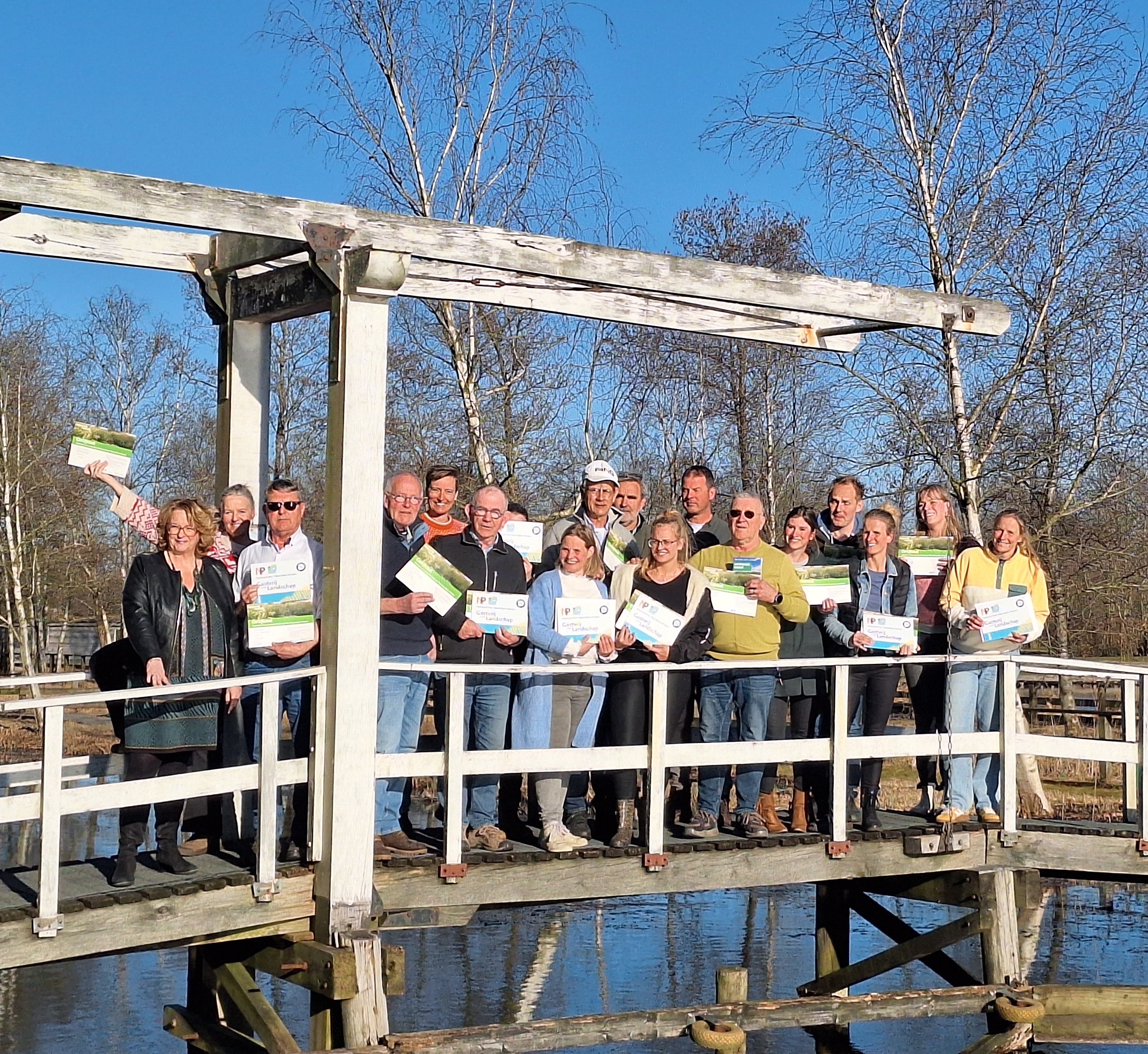 Groep mensen op een houten brug, poseren met certificaten onder een heldere, blauwe lucht.