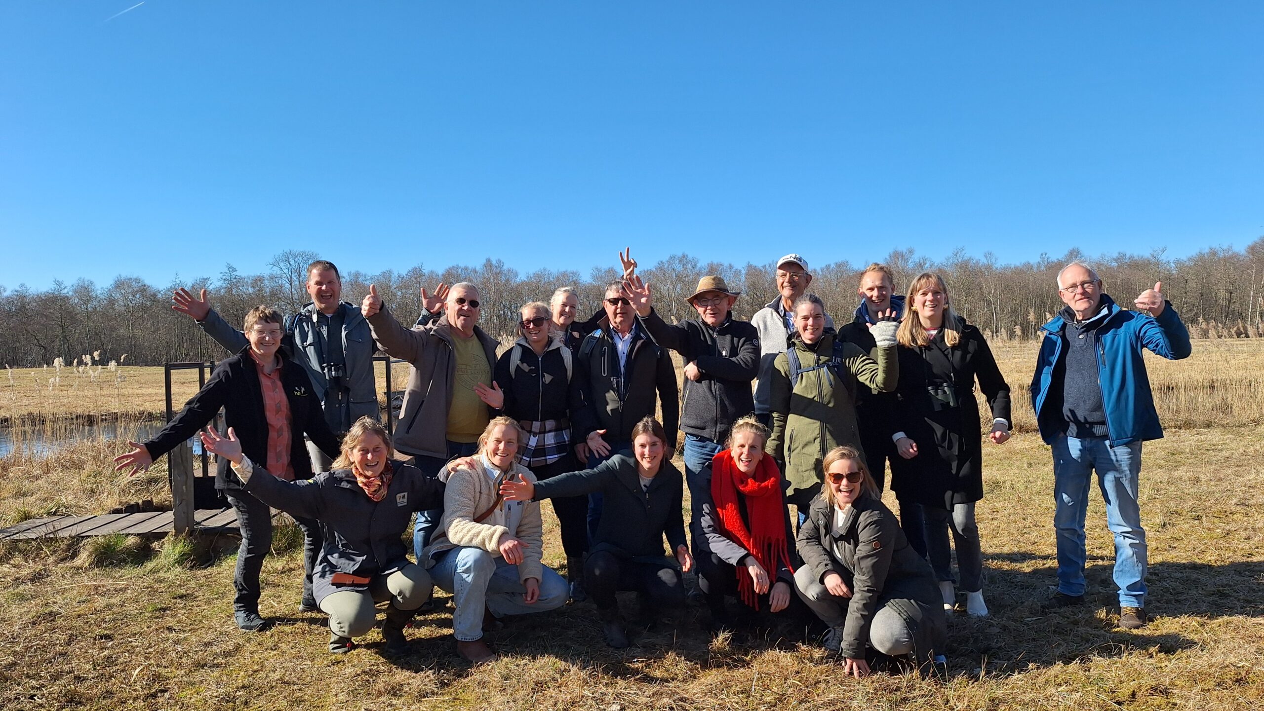 Groep mensen poseert buiten op zonnige dag, omringd door grasland en bomen.