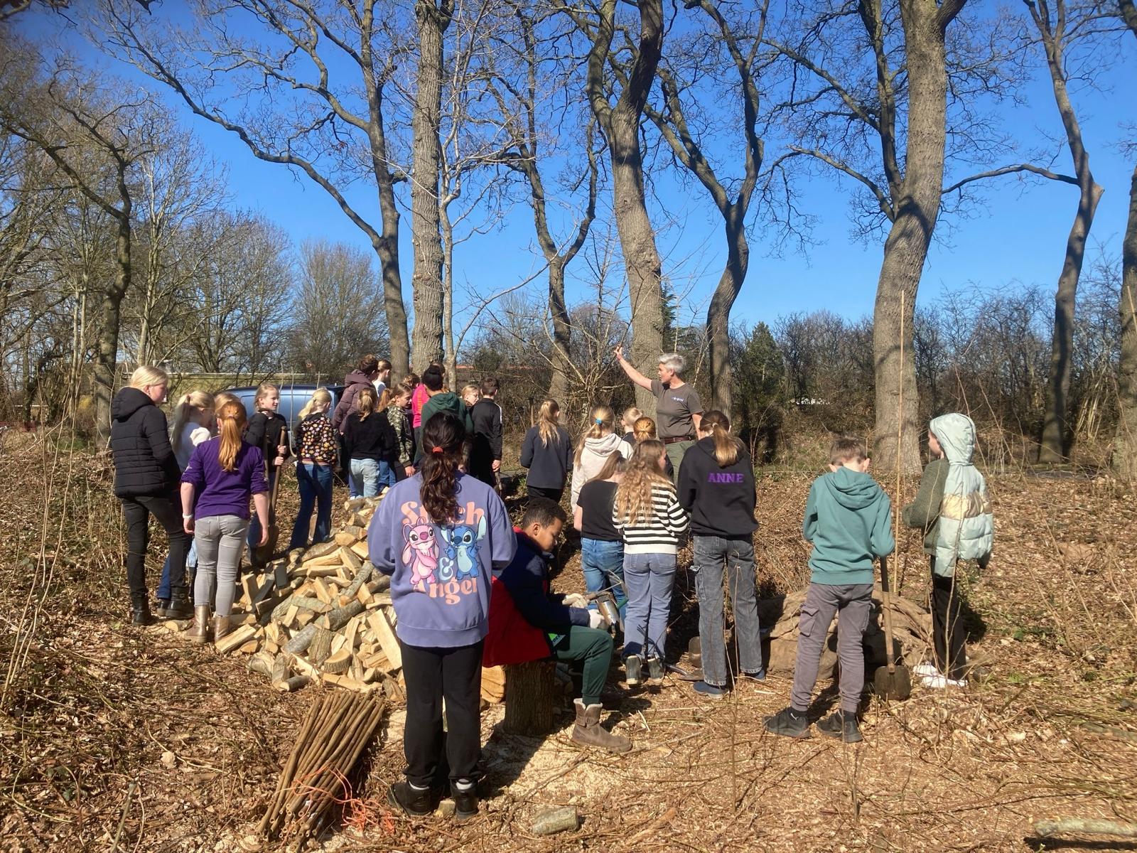 Groep kinderen luistert naar een man in een bos, omgeven door stapels hout.