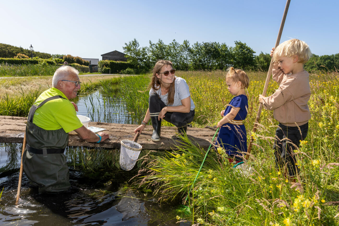 Een man vangt waterdiertjes in een sloot met een vrouw en twee kinderen op de oever.