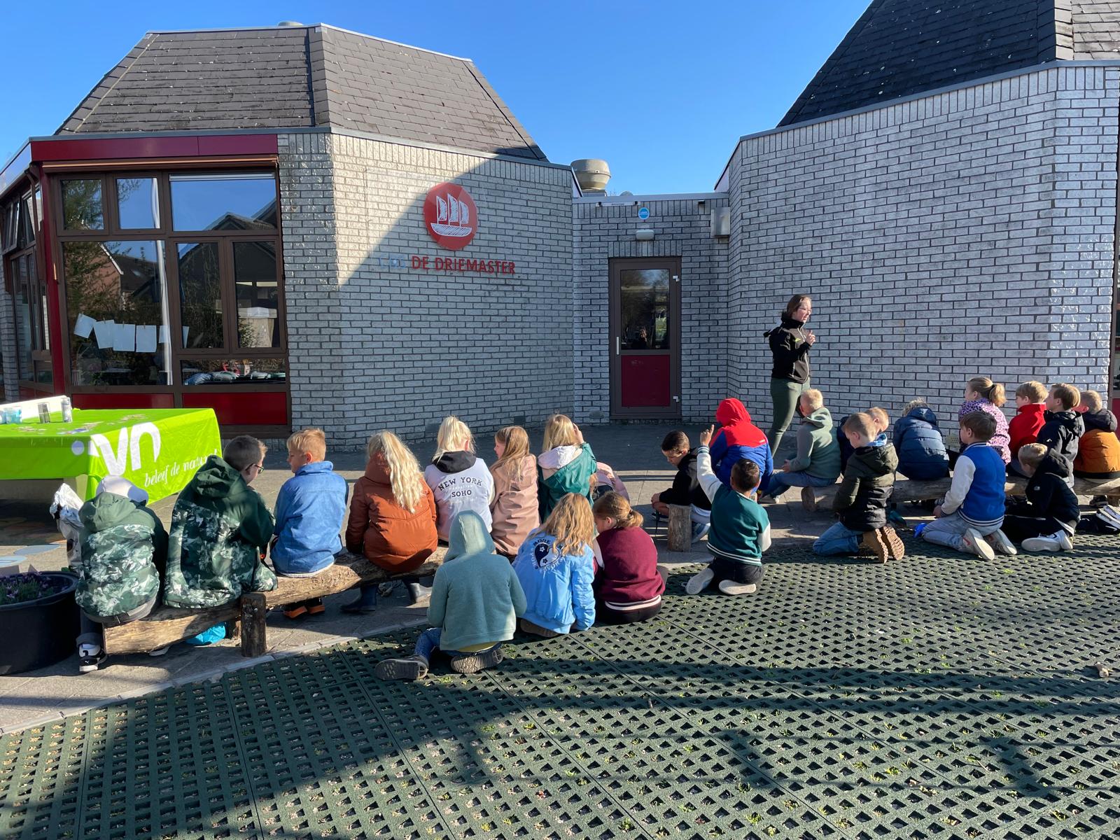 Een groep kinderen zit buiten en luistert naar een spreker bij de ingang van een schoolgebouw.