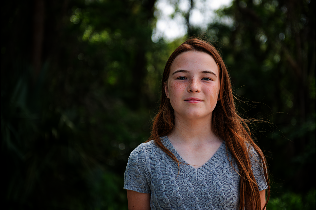 Een meisje met rood haar staat buiten, omringd door groen bos. Ze draagt een grijze trui.