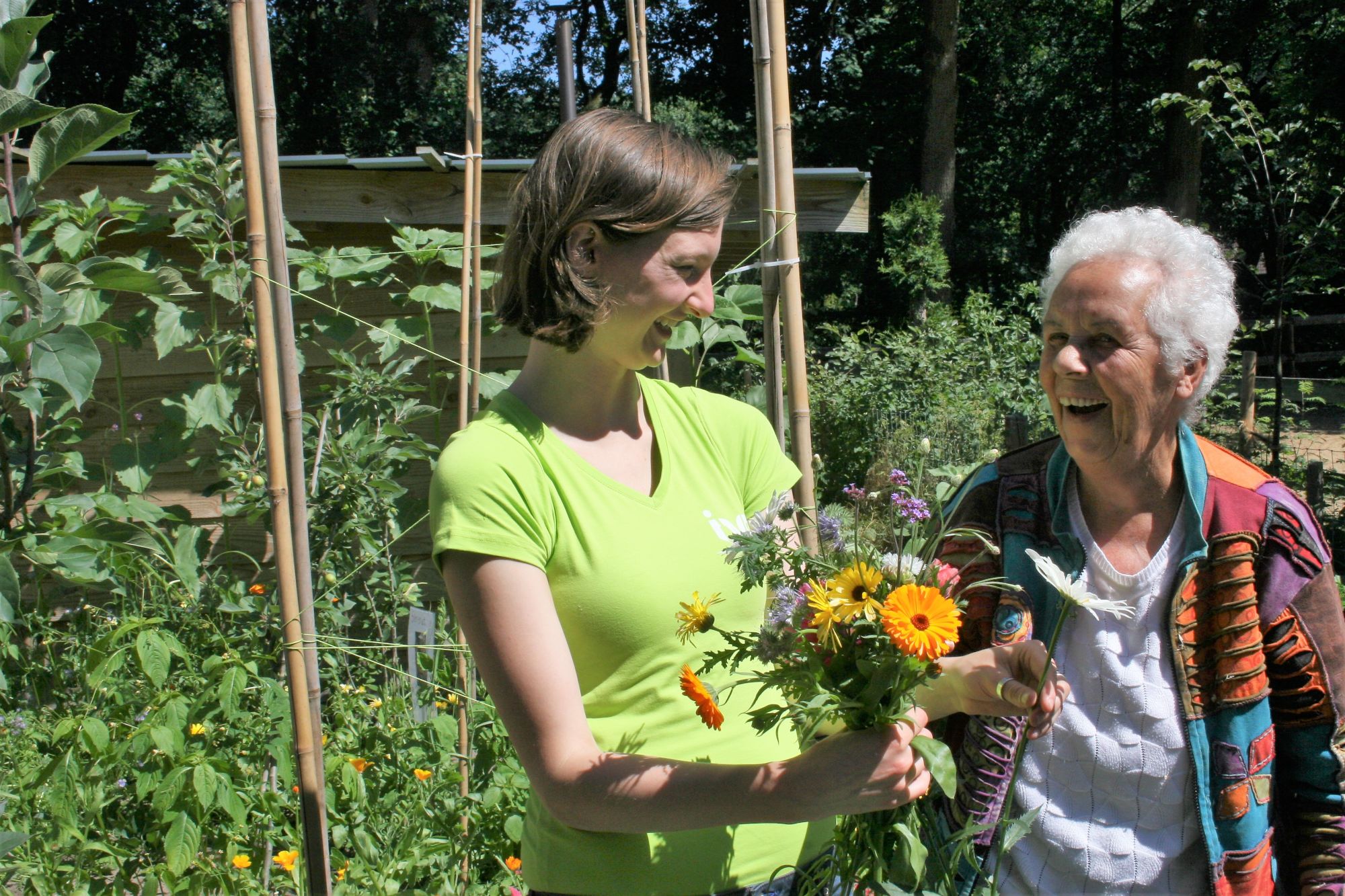 Twee vrouwen lachen en houden een boeket bloemen in een groene tuin.