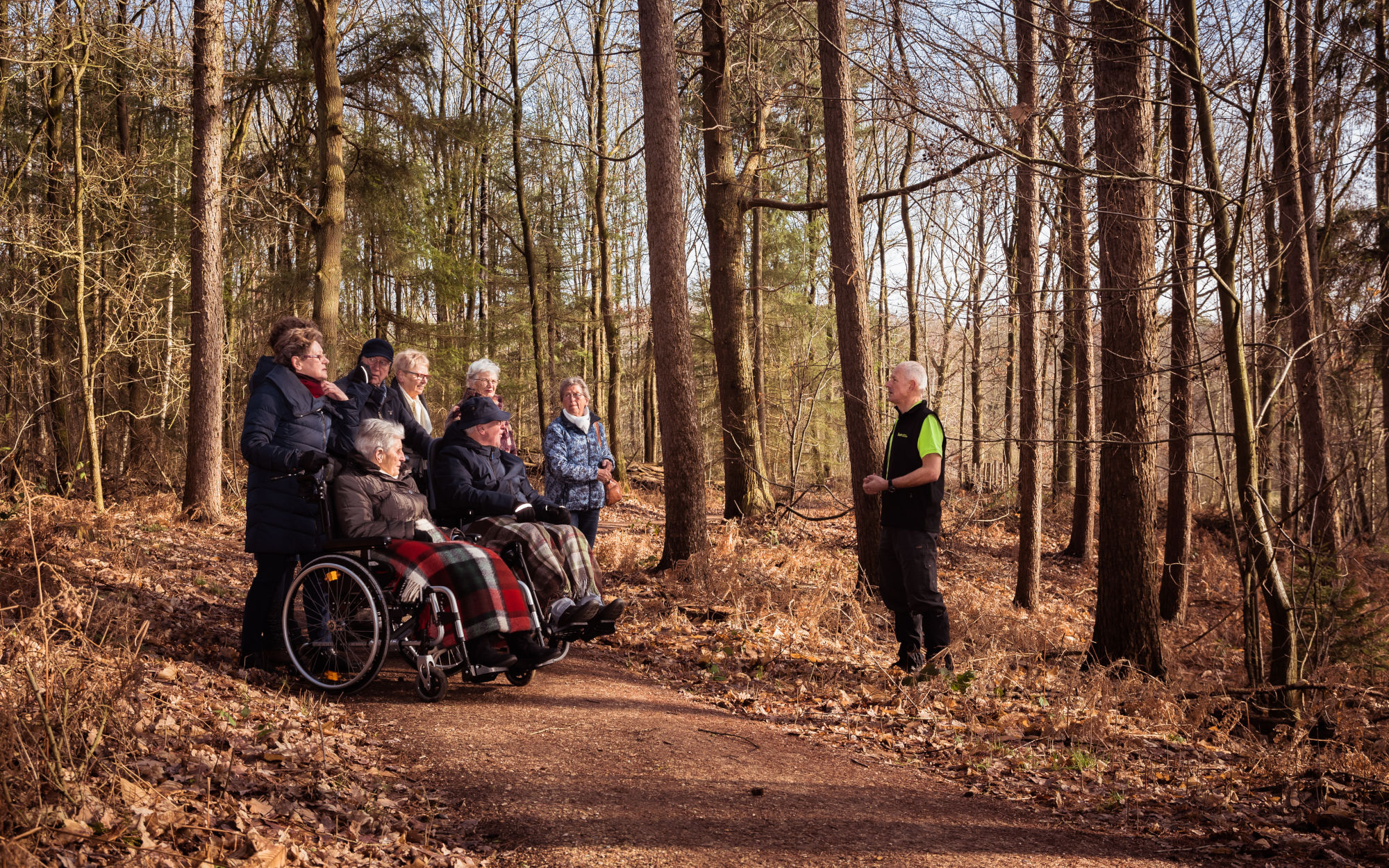 Groep ouderen in het bos met een gids, twee mensen in rolstoelen op een bospad.