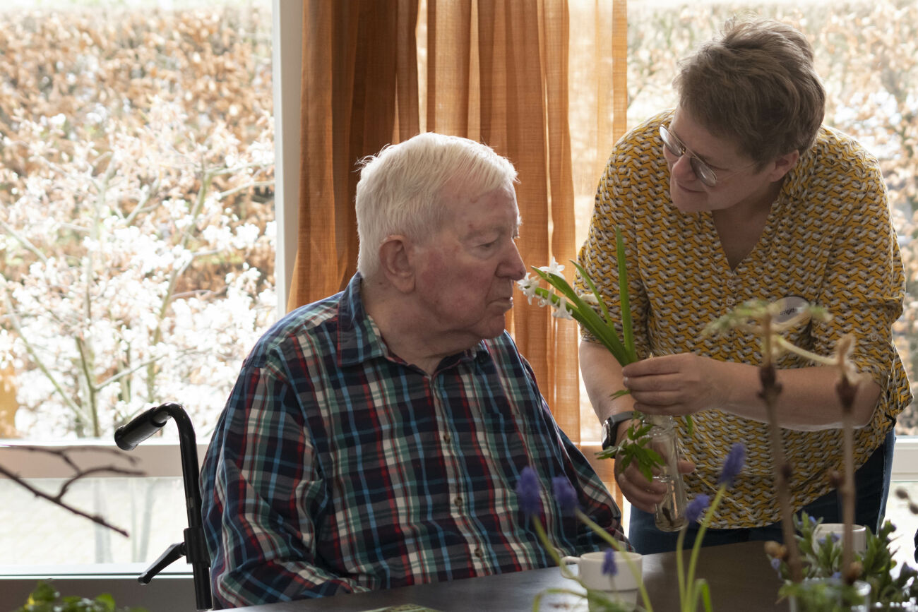 Oudere man ruikt een bloem, aangereikt door een vrouw in een huiselijke omgeving.