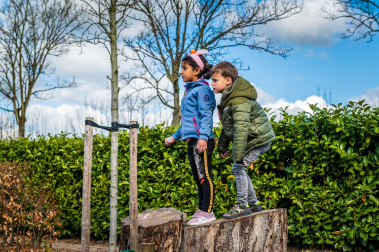 Twee kinderen staan op boomstammen in een groene tuin; bomen en wolken op de achtergrond.