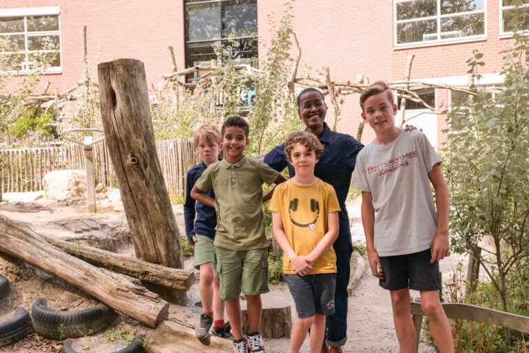 Een groep kinderen en een volwassene poseren buiten bij een schoolmuur en houten speeltoestellen.