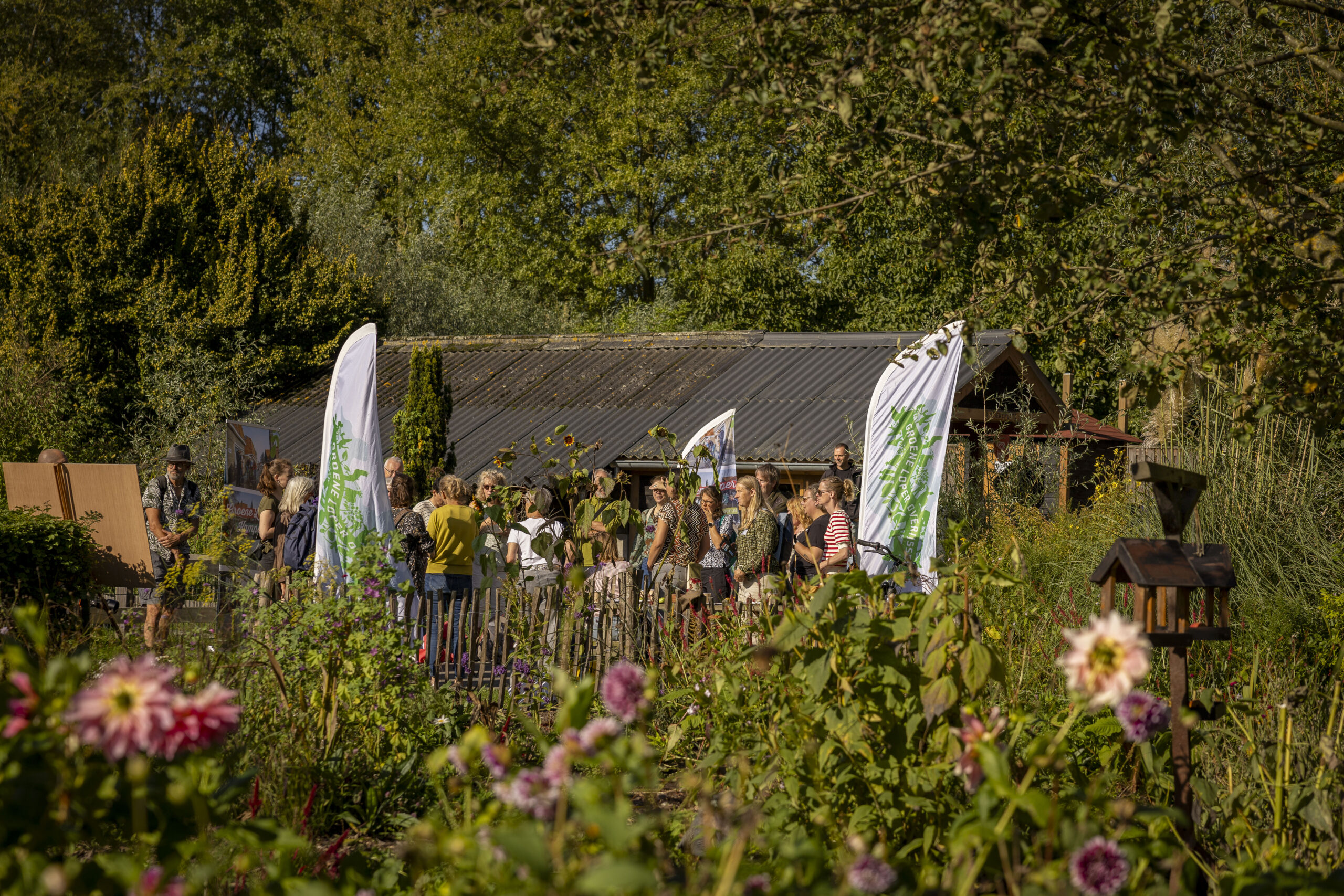 Groep mensen verzameld in een bloementuin voor een gebouw, omringd door groen en kleurrijke bloemen.
