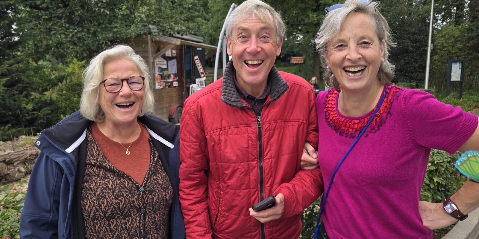 Drie lachende mensen poseren buiten voor een houten gebouw met omliggende bomen.