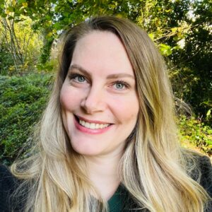 Vrouw glimlacht buiten te midden van groene bladeren en zonlicht in de achtergrond.