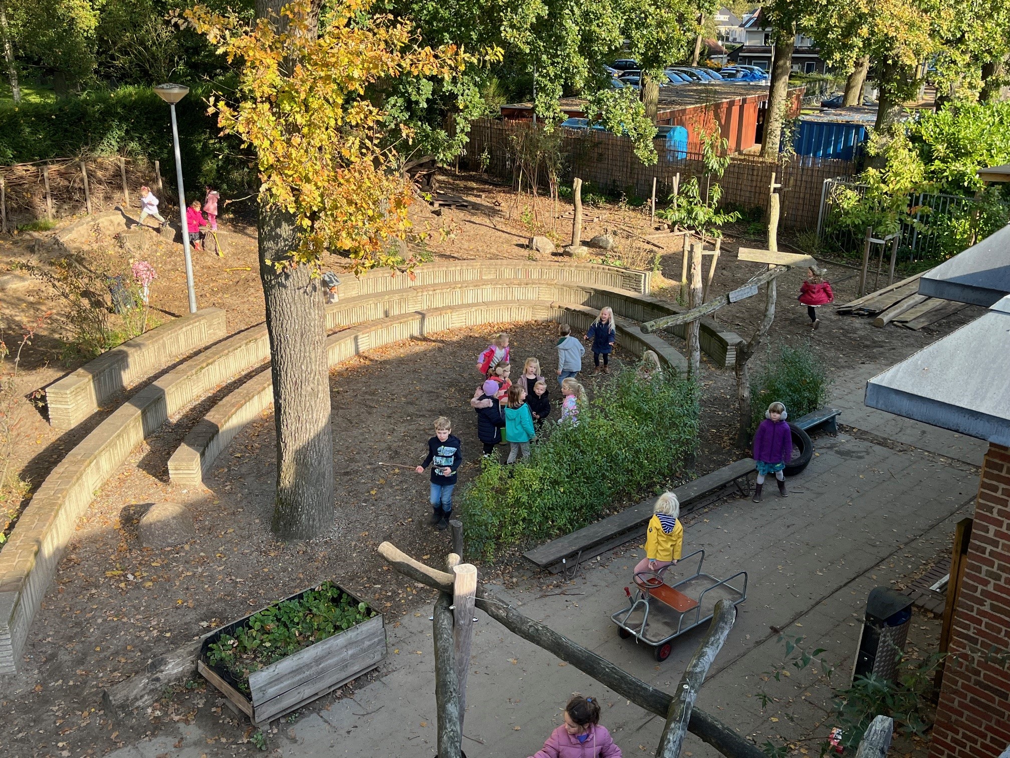 Kinderen spelen buiten op een schoolplein met bomen en zitjes rondom.