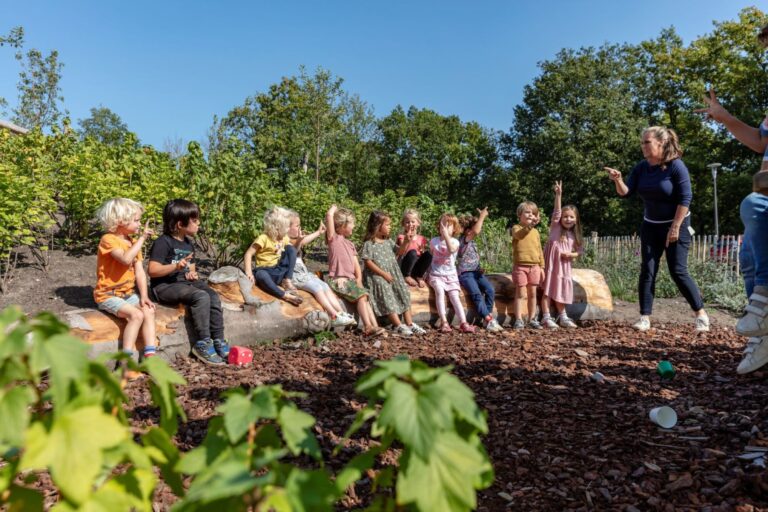 Kinderen zitten op een boomstam in een tuin, terwijl een volwassene gebaren maakt.