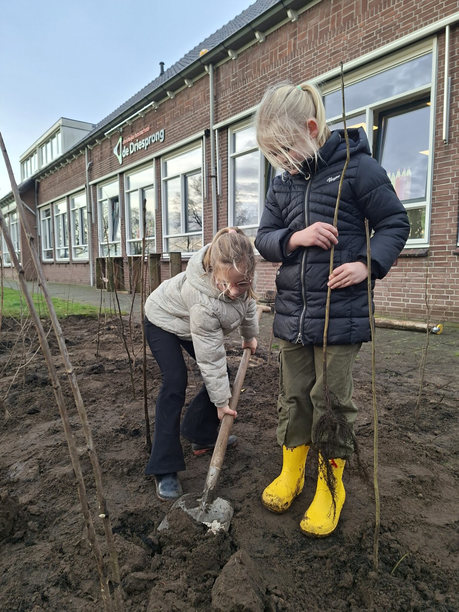 Twee kinderen planten boompjes voor een school, met een schop in modderige grond.