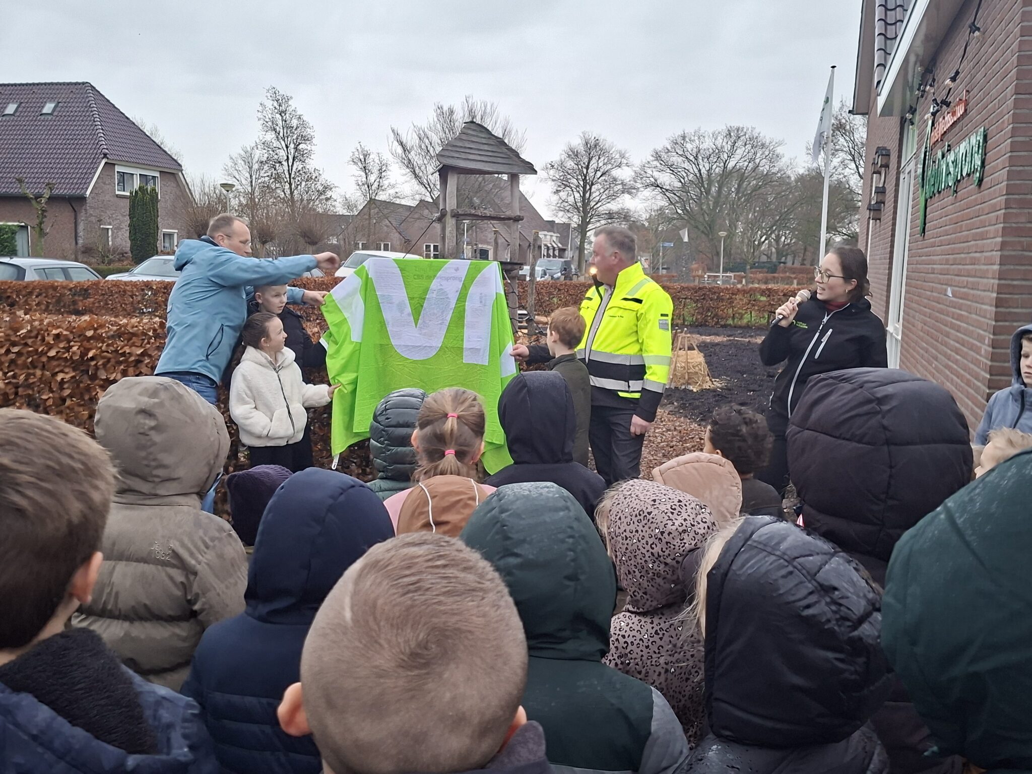 Een groep mensen verzameld bij een onthulling met een groene doek buiten bij een gebouw.