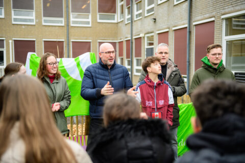 Groep mensen buiten voor een schoolgebouw, iemand spreekt enthousiast; groene vlag op de achtergrond.